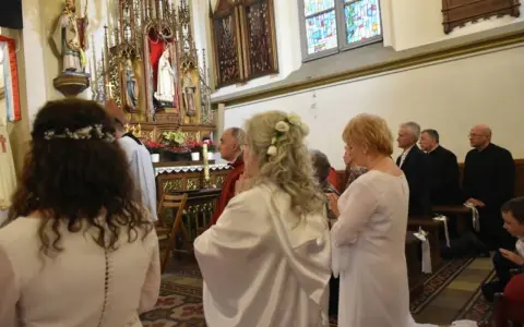 Uroczystość w kościele. Kobieta w białej sukni, z kwiatami we włosach, klęczy. To moment konsekracji, ważny dla dziewicy konsekrowanej.