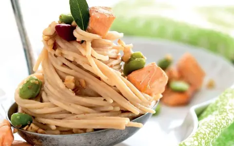Spaghetti di riso con salmone, fave e noci, guarniti con una foglia verde. Un piatto invitante e colorato.