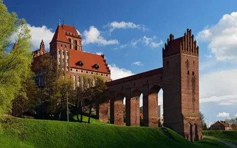 Majestatyczny zamek w Kwidzynie, z czerwonej cegły, z charakterystycznym gdaniskiem i długim, arkadowym murem obronnym, wznosi się na zielonym wzgórzu pod błękitnym niebem.