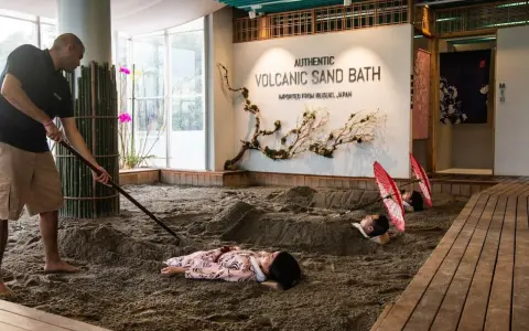 A man rakes sand for a volcanic sand bath. Two people lie buried in the sand, with red umbrellas over their heads.