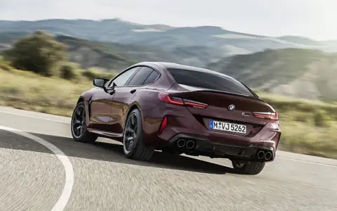 Ein dunkelroter BMW M8 Gran Coupé rast auf einer kurvigen Straße durch eine hügelige Landschaft.