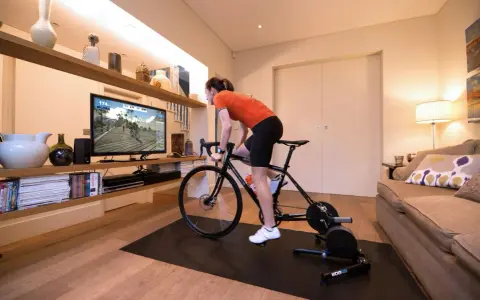 Woman in her home cycling pain cave, training indoors on a bike trainer while watching a virtual race on a large screen.