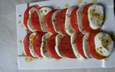 Mozzarella z pomidorami, ułożona naprzemiennie na białym talerzu, skropiona oliwą i ziołami.