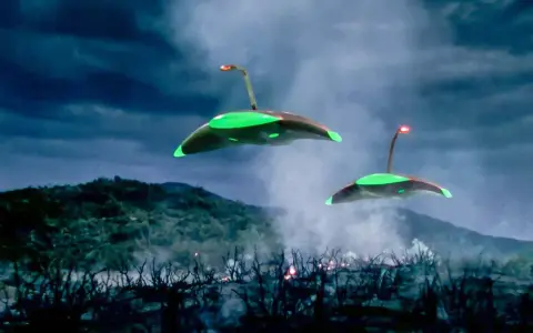 Due astronavi aliene sorvolano un paesaggio devastato, con fumo e fiamme che si levano dal terreno. La scena evoca la minaccia de "la guerra dei mondi (film 1953)".