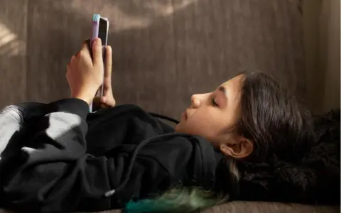 Ein Mädchen liegt auf einer Couch und schaut auf ihr Smartphone. Sie ist in ein schwarzes Kapuzensweatshirt gekleidet. Dies spiegelt aktuelle Trends der Jugend wider.