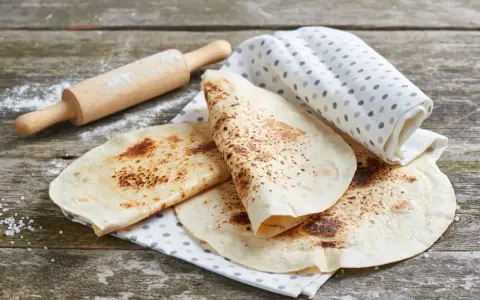 Świeżo usmażona tortilla z patelni, lekko przypieczona, leży na lnianej ściereczce obok wałka.