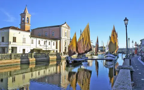 Barche a vela tradizionali ormeggiate sul canale di Cesenatico mare, con edifici storici e riflessi sull'acqua.