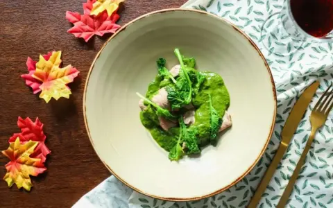 Piatto di carne con salsa verde cremosa, guarnito con cime di rapa fresche. Accanto, foglie autunnali e posate dorate.