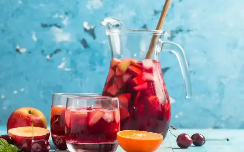 En karaff med röd bål, fylld med frukt och is, står bredvid två glas. Äpplen, körsbär och en apelsinskiva dekorerar bordet. Ett perfekt bål recept för sommaren.