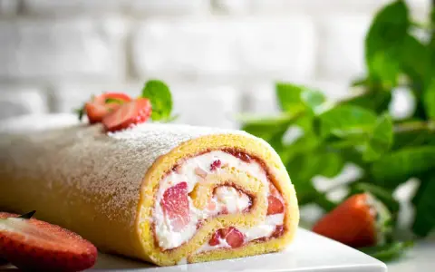 Delizioso rotolo panna e fragole, spolverato di zucchero a velo, con fragole fresche e menta sullo sfondo.