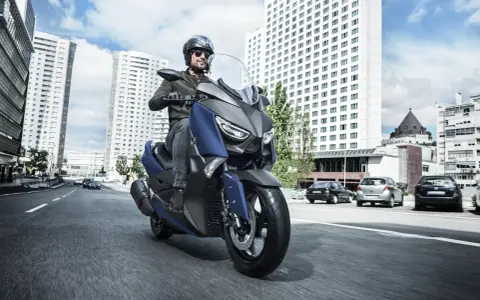 Mężczyzna w kasku i okularach przeciwsłonecznych jedzie niebieskim skuterem Yamaha X-MAX 400 po miejskiej drodze. W tle wysokie budynki i samochody.