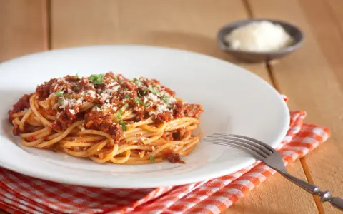 Talerz spaghetti bolognese z parmezanem i natką pietruszki, obok widelec i ser w miseczce.