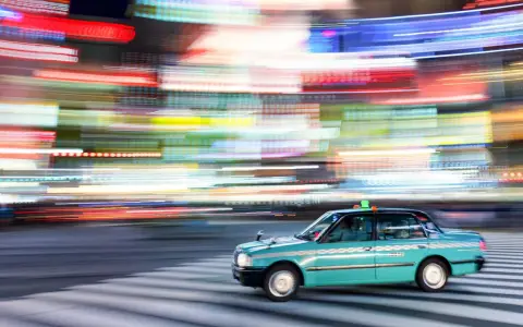 Un taxi azzurro sfreccia su strisce pedonali, con luci sfocate di città che creano un effetto dinamico, perfetto per imparare come fare foto in movimento.
