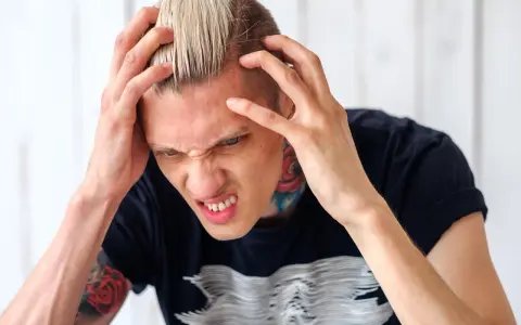 Mężczyzna z blond włosami i tatuażami na szyi i ramionach, zaciśnięte zęby i ręce na głowie, cierpi na klasterowy ból głowy.
