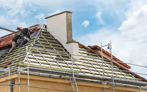 Deux couvreurs travaillent sur une toiture en cours de renovation. Des échafaudages entourent la maison.