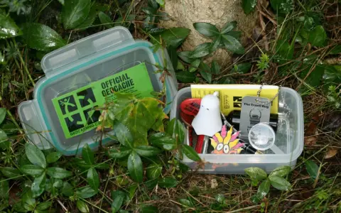 Dwa pojemniki z zawartością do geocaching co to. W jednym zielona naklejka "Official Geocache", w drugim drobne przedmioty jak duch, lupa i "Travel Bug".