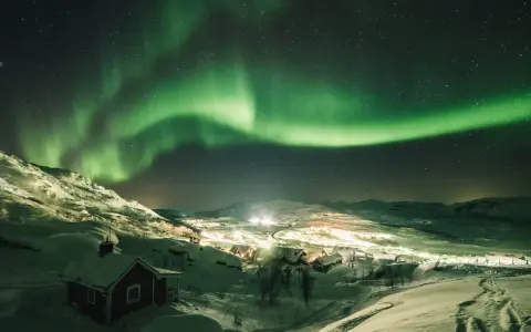 Snöklädda berg och en liten by badar i norrskenets gröna sken. En magisk natt att se norrsken.