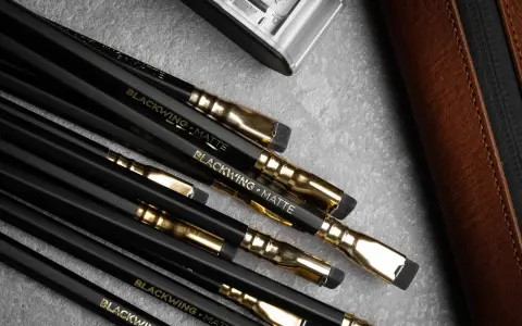 En samling svarta Blackwing-pennor med guldfattningar och suddgummin. En brun läderfodral och en metallbehållare syns också.