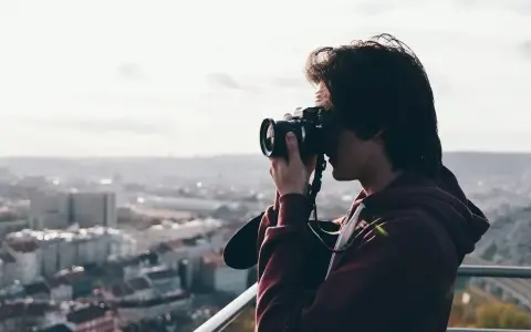 Młody mężczyzna z aparatem w rękach, uchwycony w kadrze z widokiem na miasto. Jego wzrok skupiony jest na obiektywie, jakby chciał uwiecznić piękno chwili za pomocą viewnx 2.