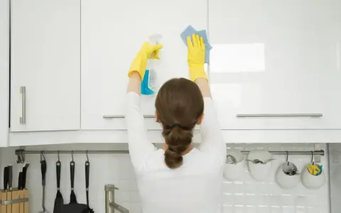 Mujer con guantes amarillos realiza limpieza armarios cocina, rociando y limpiando con paño azul.