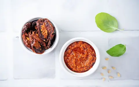 Due ciotoline bianche: una con pomodori secchi interi, l'altra con un invitante pesto di pomodori secchi. Foglie di basilico e pinoli completano la scena.
