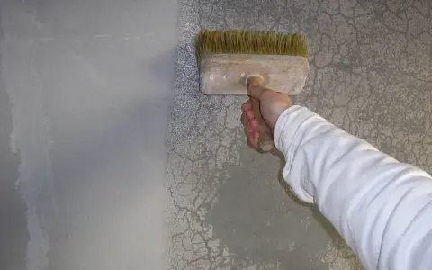 Ręka w białej rękawie nakłada szpachlę na pęknięcia na szarej ścianie za pomocą pędzla.