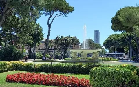 Fontana z wodą w **Parco Fellini Rimini**, otoczona zielenią i kwiatami. W tle widać autobus i wieżowiec.