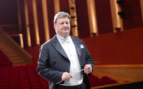 Jacek Boniecki, dyrygent w eleganckim fraku, z batutą w dłoni, na tle czerwonej widowni. Jego życiorys to historia pasji do muzyki.