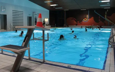 Menschen schwimmen im **Gartenhallenbad Enger**. Ein Sprungbrett mit einem orange-weißen Kegel steht am Beckenrand.