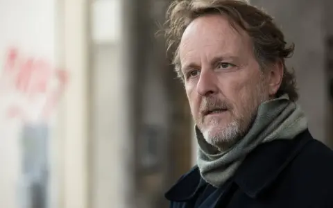 Christian Peppard, con un foulard e un cappotto scuro, guarda pensieroso verso destra.