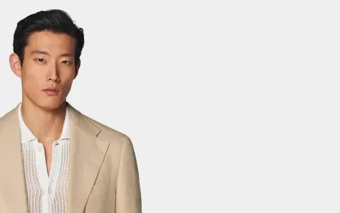A man models a tan linen blazer over a white textured polo shirt, perfect for a smart casual dress code for a wedding.