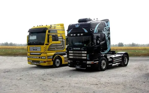 Dwa ciągniki siodłowe: żółty MAN i czarna Scania z grafiką. Różne rodzaje naczep czekają na załadunek.