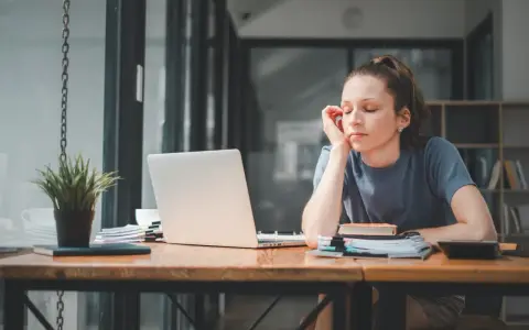 Zmęczona dziewczyna przy biurku, zamyślona, jakby zastanawiała się, ostracyzm co to. Laptop, książki i roślina na blacie.