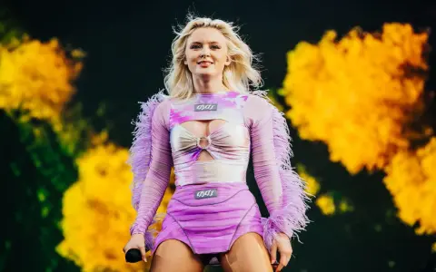 Zara Larsson konsert. Hon står på scenen i en lila och rosa outfit med fjädrar på ärmarna.