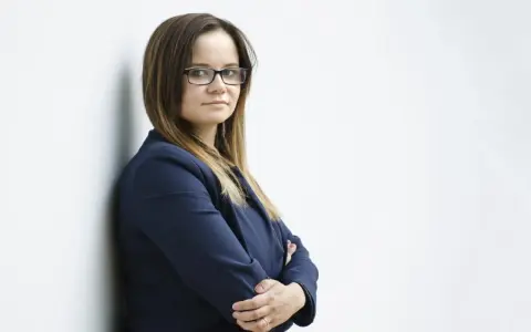 Kobieta w okularach i granatowej marynarce stoi oparta o ścianę, ze skrzyżowanymi rękami.