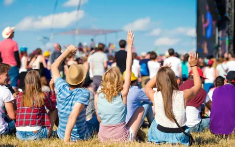 Widok zza pleców na tłum ludzi bawiących się na jednym z letnich festiwali w Polsce. Słońce świeci, niebo jest błękitne.