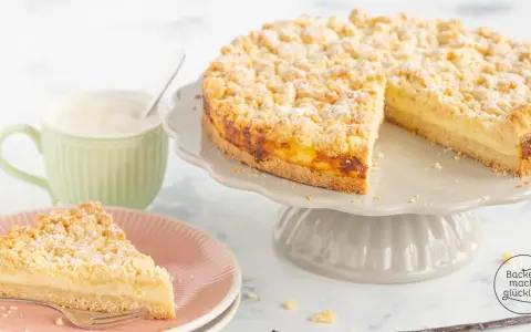 Ein saftiger Streuselkuchen mit Puddingfüllung, bestäubt mit Puderzucker, neben einer Tasse Cappuccino.