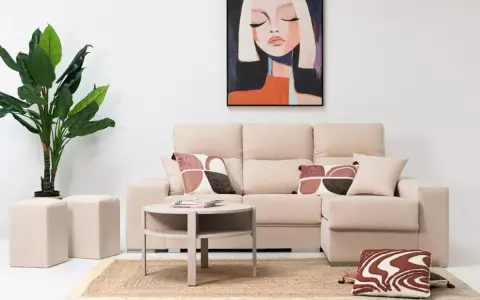 Sofá modular beige con chaise longue a la derecha, mesa auxiliar redonda, alfombra de yute, plantas y cuadro abstracto.