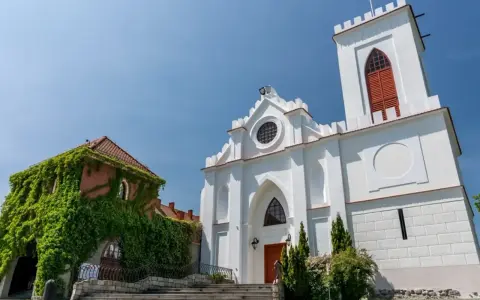 Biały kościół z wieżą i zarośnięty bluszczem budynek na tle błękitnego nieba.