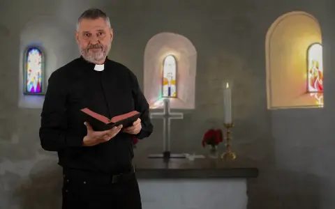 Ein Mann mit grauem Bart und Priesterkragen hält ein Buch in einer Kirche. Er hat sich entschieden, Priester zu werden.