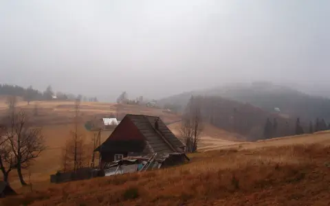Mglisty krajobraz na przełęczy Przegibek. Drewniany dom z ciemnym dachem, otoczony suchą trawą i drzewami.