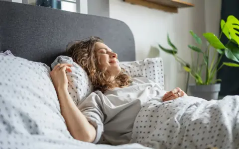 Une femme souriante se repose dans un lit douillet. Elle semble heureuse, une belle illustration de comment choisir une bonne literie pour un sommeil réparateur.