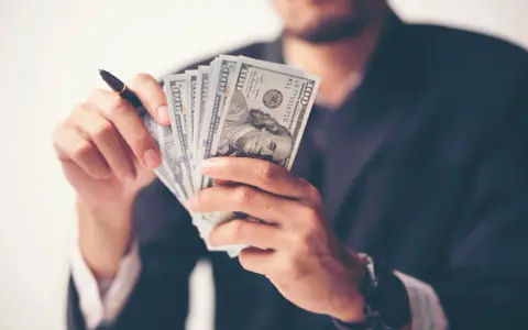 Mężczyzna w garniturze trzyma plik banknotów 100 USD. To wizualizacja rachunku przepływów pieniężnych krok po kroku.