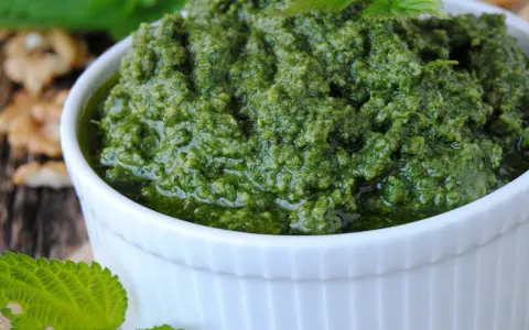 Aksamitne, zielone pesto z pokrzywy w białej miseczce, obok orzechy włoskie i liście pokrzywy.