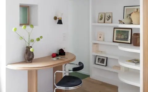 Un despacho pequeño en casa con un escritorio curvo de madera, una silla de terciopelo azul y estanterías blancas llenas de libros y objetos decorativos.
