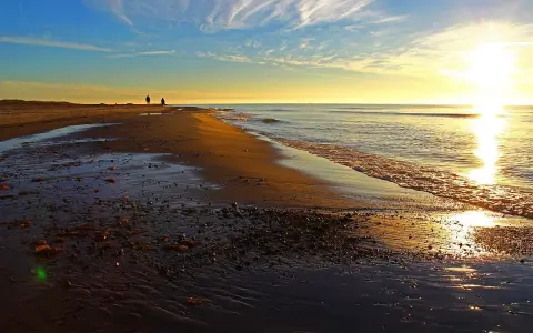 Två personer promenerar längs stranden vid Skagens udde i solnedgången. Vågorna sköljer in mot land.