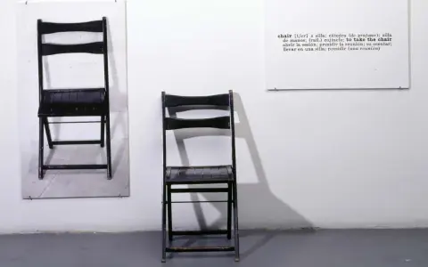 Una y tres sillas. Dos sillas negras plegables, una colgada en la pared y otra de pie, junto a un cartel con la definición de "chair".