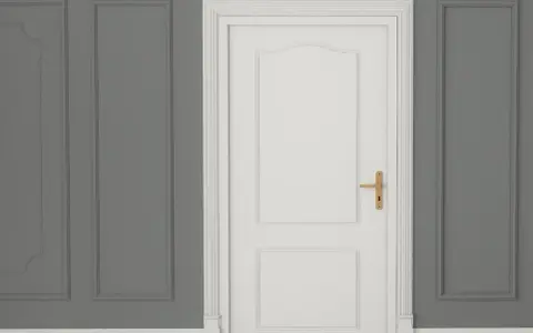 Białe drzwi z ozdobną listwą opaskową do drzwi i złotą klamką, na tle szarej ściany z panelami.