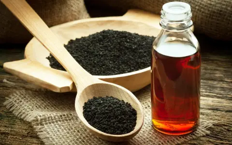 Huile de nigelle ambrée dans un flacon, à côté de graines noires dans une cuillère en bois. Découvrez les bienfaits de l'huile de nigelle.