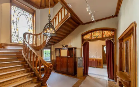 Escalier majestueux en bois, vitraux colorés, mobilier ancien. Un intérieur d'une élégance intemporelle, évoquant le style Majorelle.